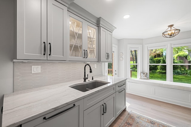 kitchen featuring light stone counters, a sink, light wood-type flooring, backsplash, and glass insert cabinets
