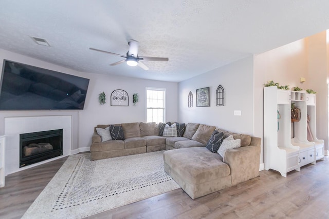 living area featuring a ceiling fan, a textured ceiling, visible vents, wood finished floors, and a glass covered fireplace