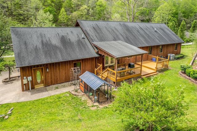 rear view of property with a wooden deck, a lawn, a shingled roof, and a view of trees
