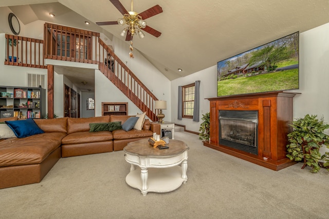 living area with carpet floors, ceiling fan, stairway, a glass covered fireplace, and high vaulted ceiling