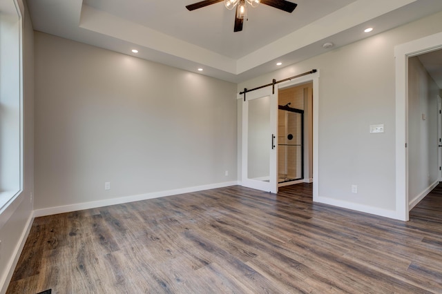 empty room with dark wood-style flooring, a barn door, a raised ceiling, a ceiling fan, and recessed lighting