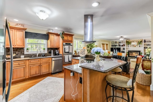 kitchen featuring a sink, stainless steel appliances, island exhaust hood, a breakfast bar, and a glass covered fireplace