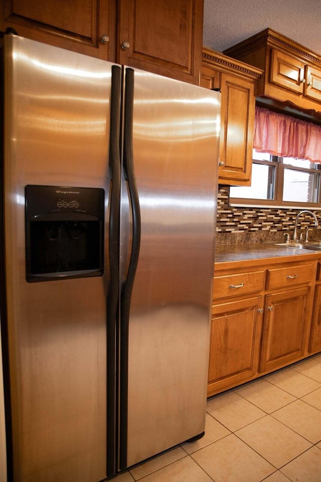 kitchen featuring light tile patterned flooring, dark countertops, stainless steel fridge, brown cabinets, and a sink