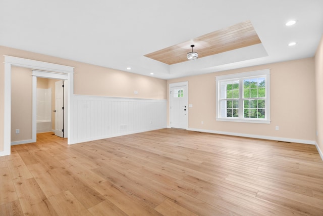 unfurnished living room featuring light wood-style flooring, a raised ceiling, a wainscoted wall, recessed lighting, and baseboards