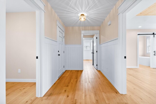corridor featuring a barn door, light wood finished floors, and a wainscoted wall