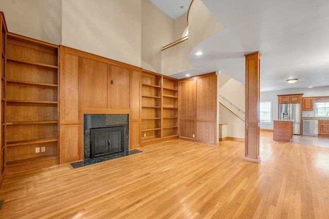unfurnished living room featuring light wood-style flooring, built in shelves, stairs, a fireplace, and a high ceiling