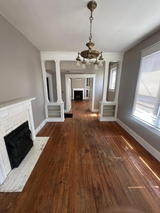 unfurnished living room featuring a brick fireplace, a chandelier, hardwood / wood-style floors, baseboards, and ornate columns