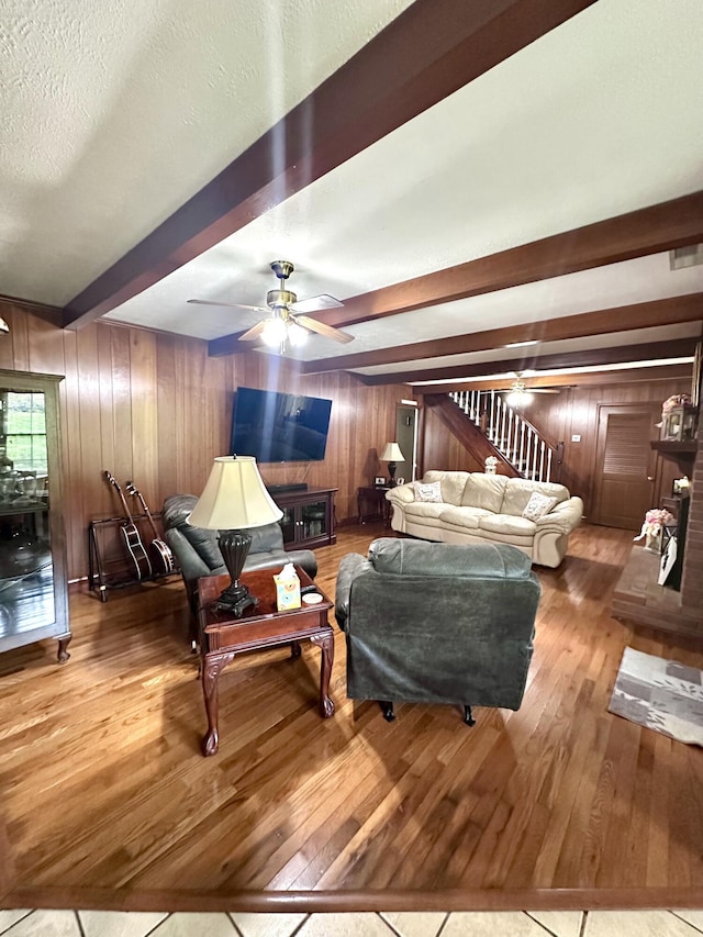 living room with a ceiling fan, beam ceiling, hardwood / wood-style floors, stairway, and wooden walls