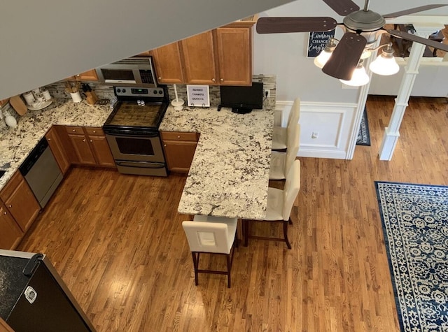 kitchen featuring wood finished floors, brown cabinets, stainless steel appliances, wainscoting, and ceiling fan
