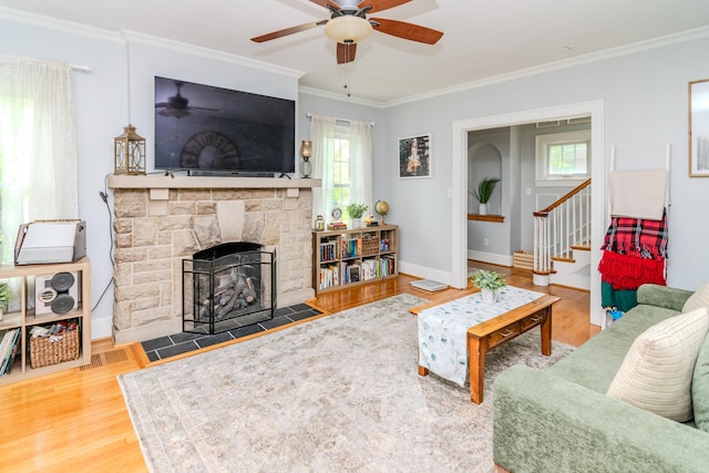 living area with ceiling fan, crown molding, a stone fireplace, wood finished floors, and stairs
