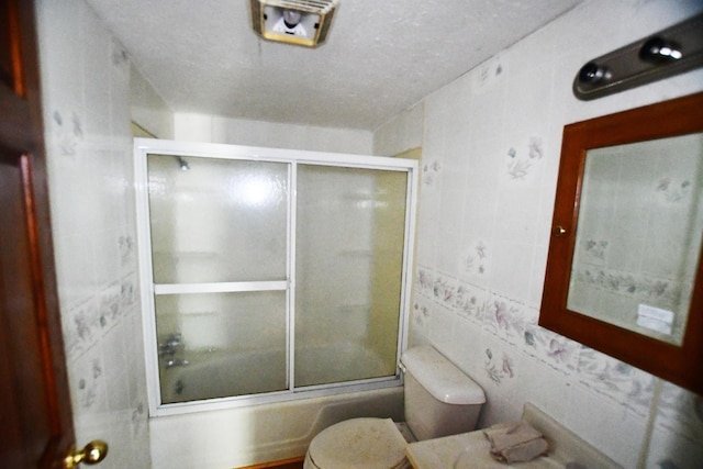 bathroom with tile walls, toilet, shower / bath combination with glass door, and a textured ceiling
