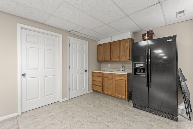 kitchen with black fridge with ice dispenser, brown cabinetry, a drop ceiling, light countertops, and baseboards