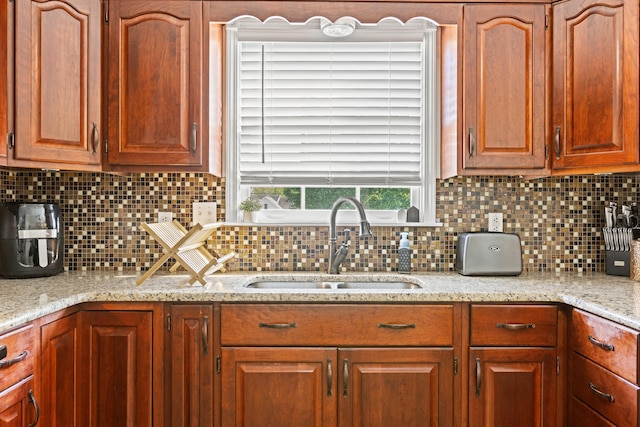 kitchen featuring a sink, tasteful backsplash, light stone countertops, and brown cabinetry