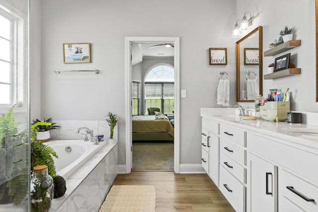 ensuite bathroom featuring wood finished floors, double vanity, a bath, ceiling fan, and baseboards