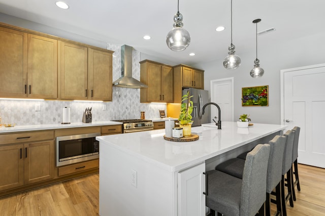 kitchen with wall chimney exhaust hood, stainless steel appliances, light wood finished floors, a sink, and a center island with sink
