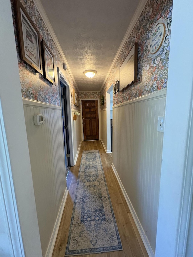 hallway featuring wallpapered walls, wainscoting, wood finished floors, ornamental molding, and baseboards