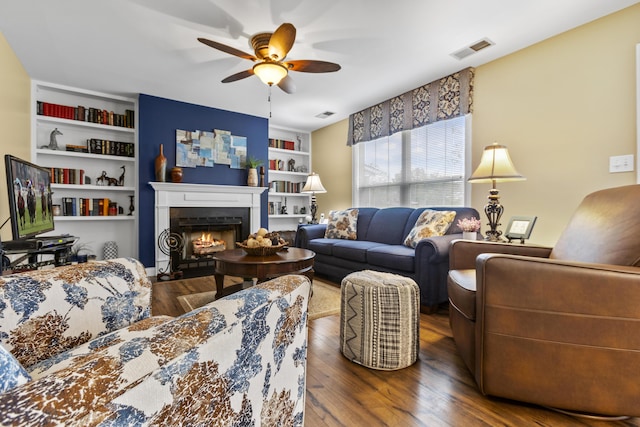 living room featuring wood finished floors, a ceiling fan, built in features, and a warm lit fireplace