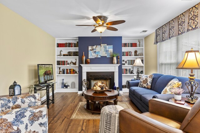 living area with ceiling fan, built in features, a warm lit fireplace, and wood finished floors