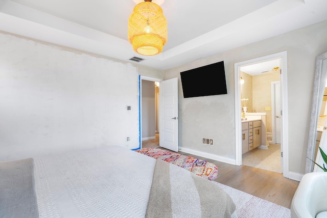 bedroom featuring light wood-type flooring, a tray ceiling, baseboards, and ensuite bath