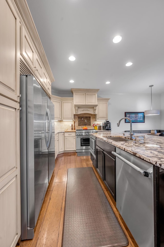 kitchen with appliances with stainless steel finishes, cream cabinetry, light wood-type flooring, a sink, and light stone countertops