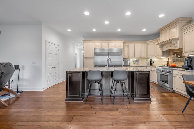 kitchen featuring cream cabinetry, high quality appliances, ornamental molding, an island with sink, and backsplash