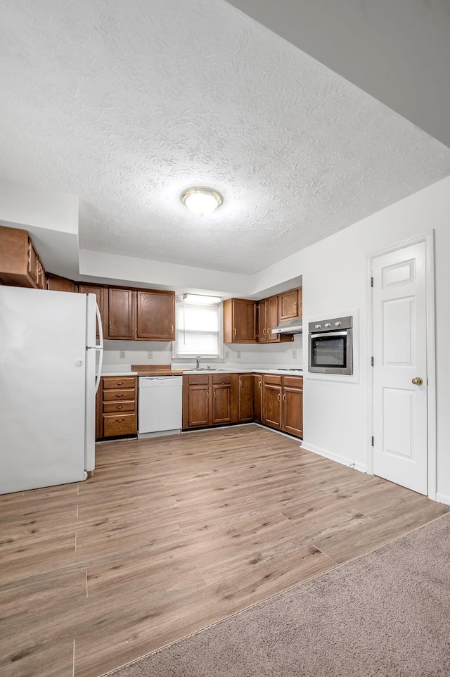 kitchen featuring white appliances, light countertops, under cabinet range hood, a textured ceiling, and brown cabinets