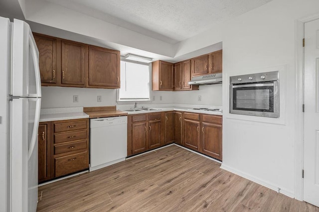 kitchen featuring white appliances, under cabinet range hood, a sink, light wood finished floors, and brown cabinets