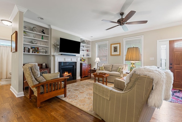 living area with ornamental molding, a lit fireplace, wood finished floors, ceiling fan, and built in shelves