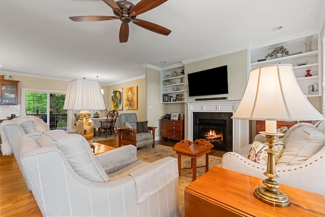 living room featuring light wood-style floors, crown molding, a lit fireplace, a ceiling fan, and built in features