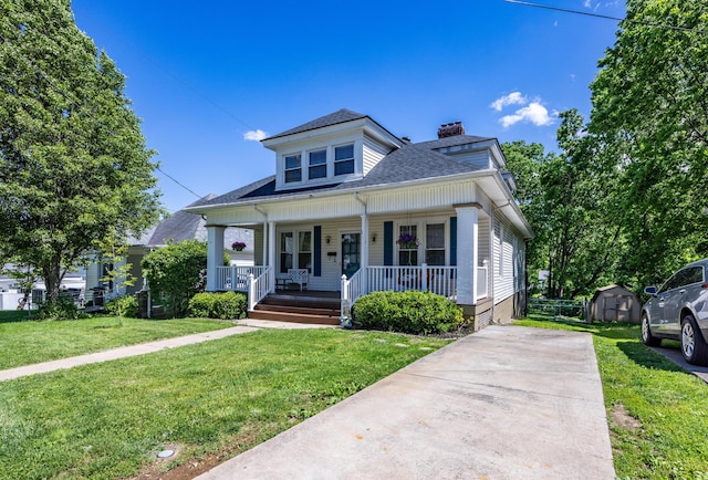 bungalow featuring a chimney, covered porch, a storage shed, a shingled roof, and a front yard
