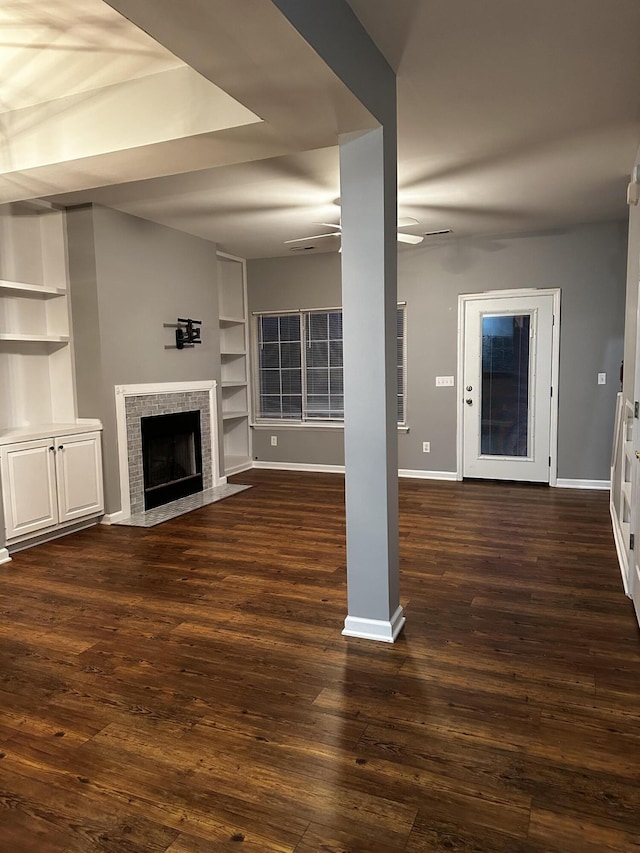 unfurnished living room featuring ceiling fan, built in shelves, a tiled fireplace, baseboards, and dark wood-style floors