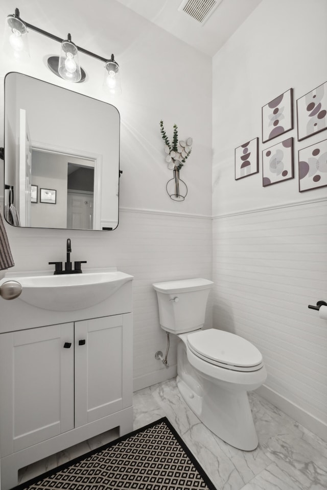 bathroom featuring marble look tile flooring, toilet, vanity, wainscoting, and baseboards