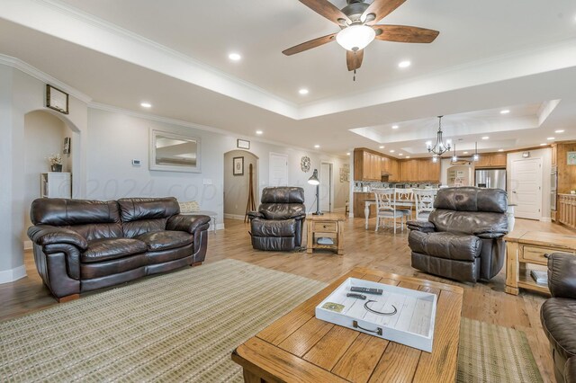 living room with light wood-type flooring, ceiling fan with notable chandelier, a raised ceiling, and ornamental molding