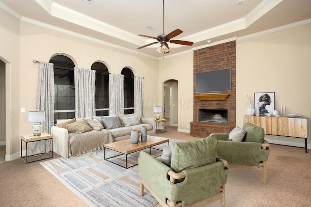living room with carpet flooring, a ceiling fan, a brick fireplace, a raised ceiling, and arched walkways