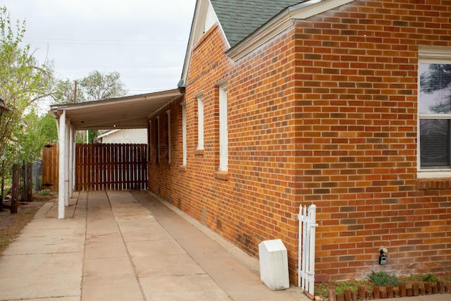 view of property exterior with driveway, a shingled roof, fence, an attached carport, and brick siding