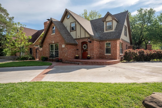 tudor home featuring brick siding, a shingled roof, a front lawn, board and batten siding, and a chimney
