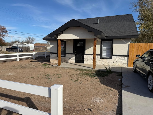 bungalow featuring stone siding, roof with shingles, fence, and stucco siding