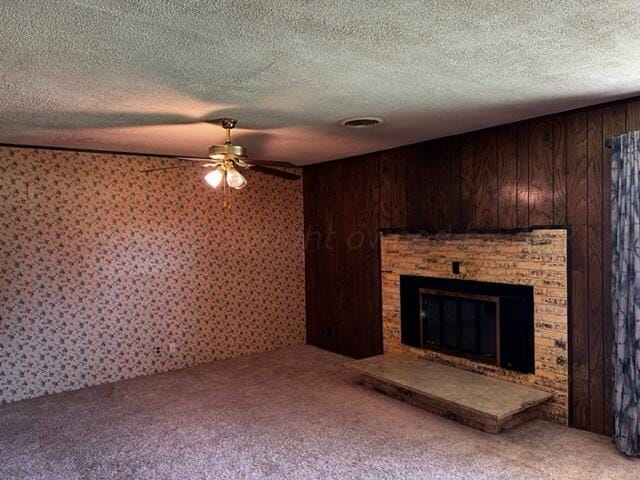 unfurnished living room with carpet, a textured ceiling, wooden walls, a brick fireplace, and ceiling fan