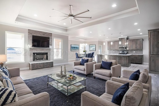 living room with a stone fireplace, crown molding, light wood-type flooring, a tray ceiling, and ceiling fan