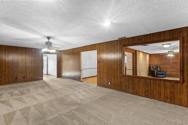 empty room with a textured ceiling, light colored carpet, ceiling fan, and wooden walls