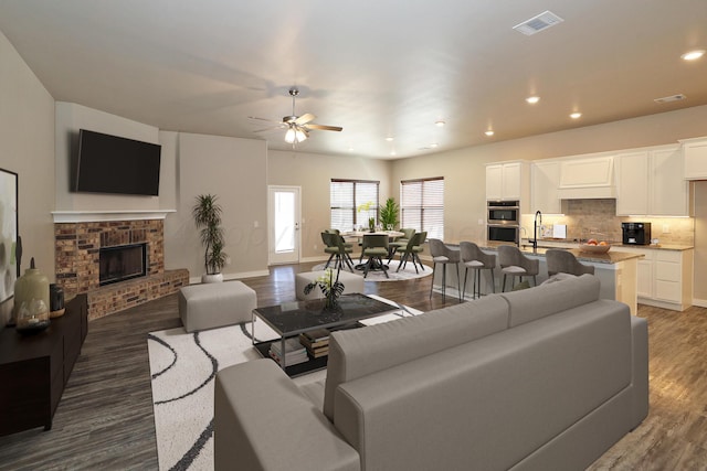 living room featuring dark wood-type flooring, ceiling fan, and a brick fireplace