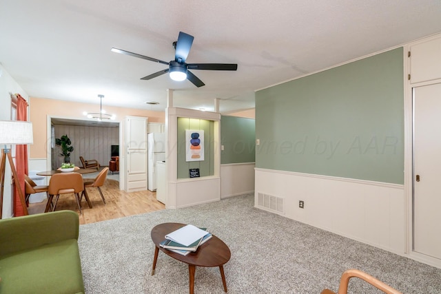 living area featuring light carpet, a wainscoted wall, ceiling fan, and visible vents