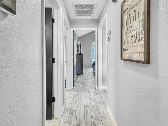 hall featuring visible vents, light wood-style flooring, baseboards, and a textured wall