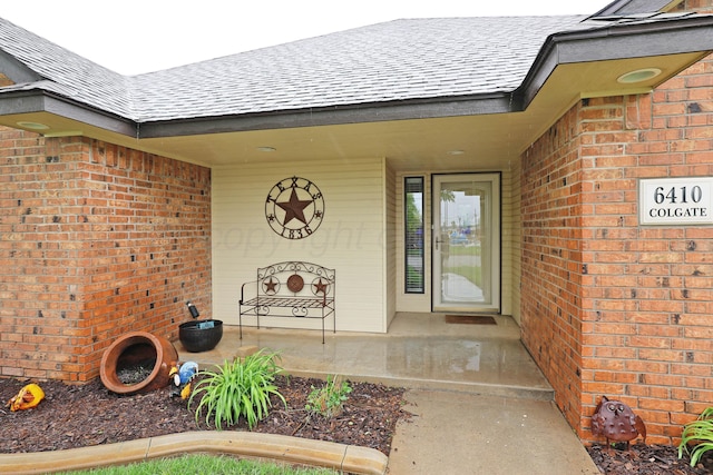 view of exterior entry with a shingled roof, brick siding, and a porch