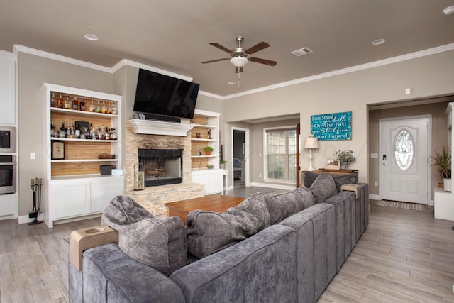 living room with a stone fireplace, crown molding, built in features, and light hardwood / wood-style flooring