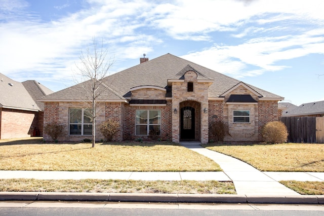 french country inspired facade with a front yard, a chimney, fence, and brick siding