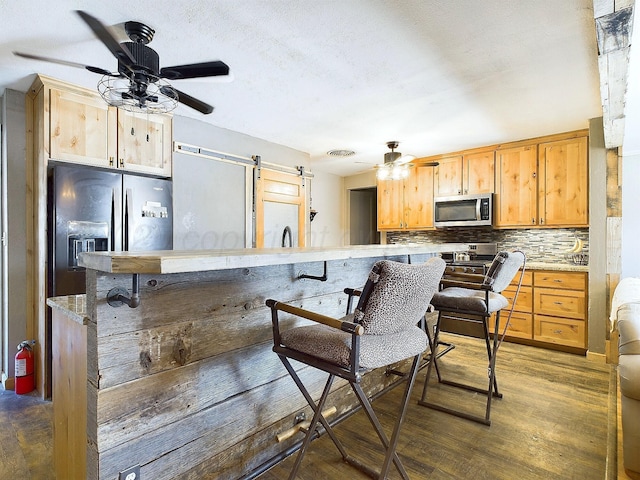 kitchen with dark wood-type flooring, ceiling fan, stainless steel appliances, decorative backsplash, and a barn door
