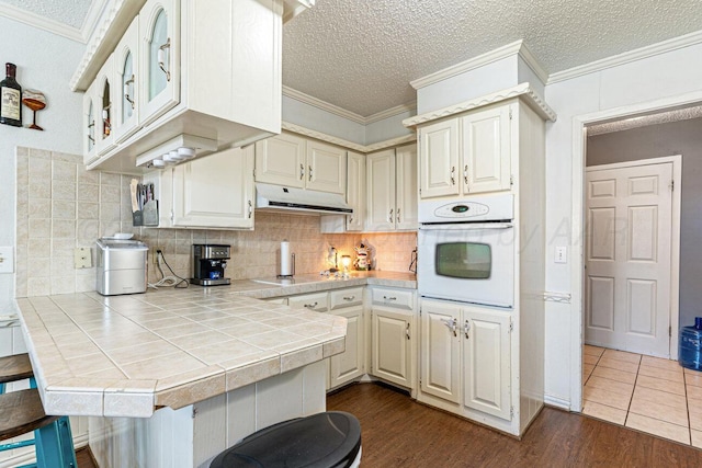 5216 Westway Trl, Amarillo, TX 79109 - kitchen featuring oven, tile counters, under cabinet range hood, ornamental molding, and dark wood-style flooring