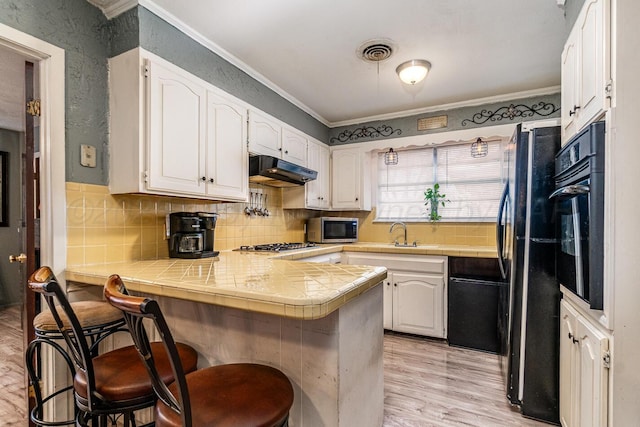 kitchen featuring visible vents, tile countertops, a peninsula, under cabinet range hood, and black appliances