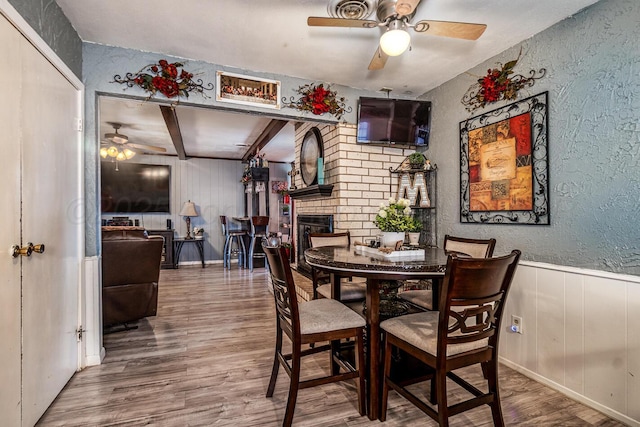 dining space featuring a ceiling fan, a textured wall, a wainscoted wall, wood finished floors, and a fireplace